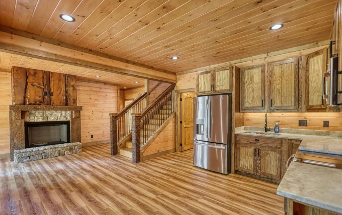 Open great room with a stone fireplace, staircase and kitchen under a pine ceiling
