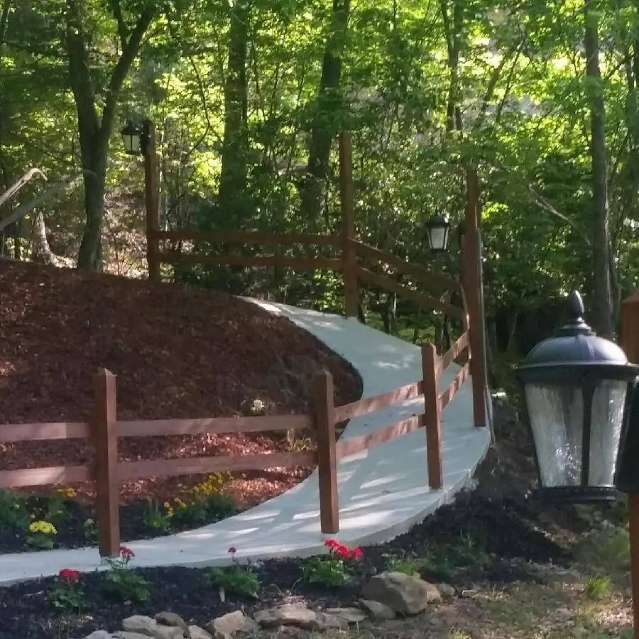 Curved concrete walkway with a stained rail fence and lantern on a wooded slope